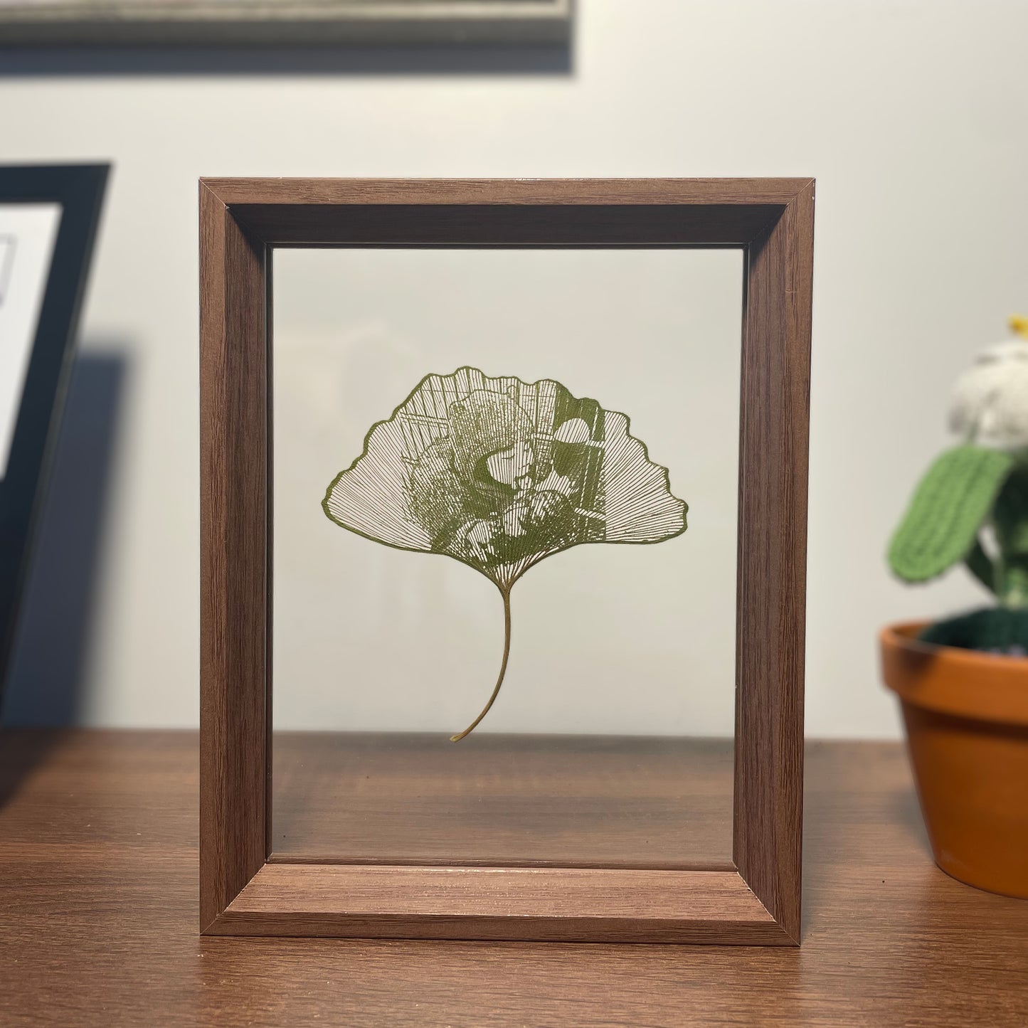 Framed artwork of a ginkgo leaf on a wooden surface with a plant in the background