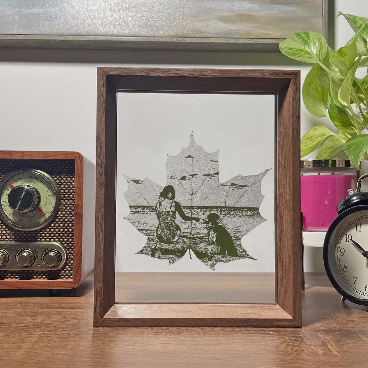 Framed artwork of a maple leaf of a woman and a dog by a lake with a plant and vintage radio in the background.