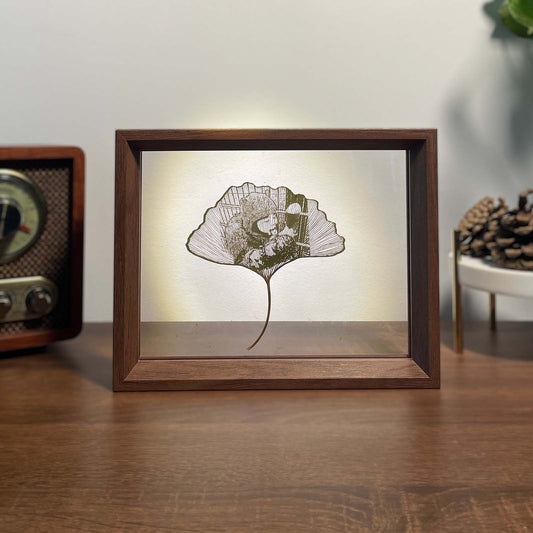 Framed artwork of a ginkgo leaf with a person inside on a wooden surface.