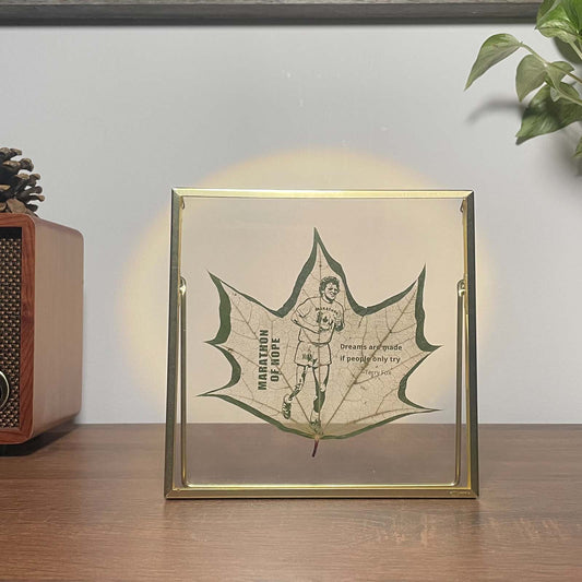 Framed artwork with a ginkgo leaf design of Terry Fox and text on a wooden surface.