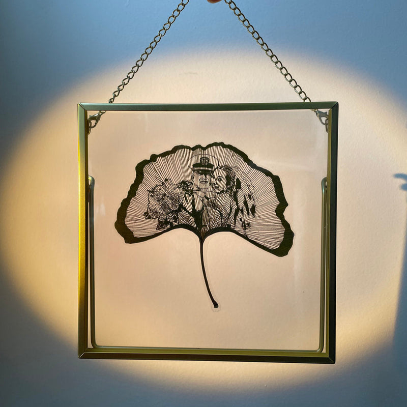 Framed artwork of a ginkgo leaf with intricate designs, held by a hand against a blue background.