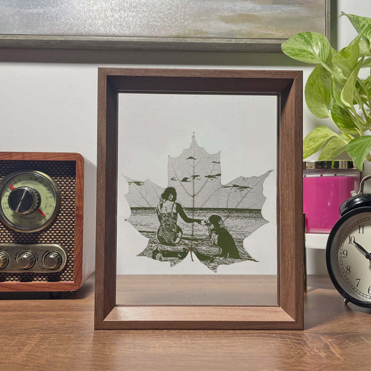 Framed artwork of a maple leaf of a woman and a dog by a lake with a plant and vintage radio in the background.