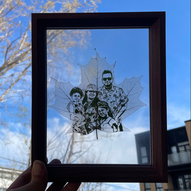 Framed artwork of a family portrait  on a maple leaf, with a blue sky and tree branches in the background