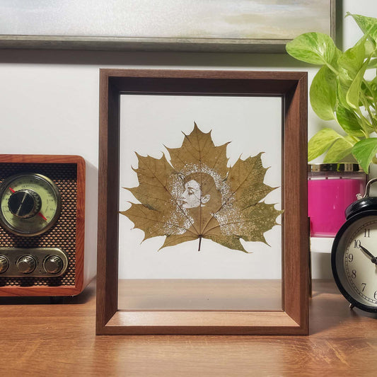 Framed artwork of a maple leaf with Audrey Hepburn's face on a desk with a vintage radio and clock.